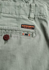 Pantaloni di tessuto verde con una finitura testurizzata, dotati di un bottone marrone, dettagli cuciti e un'etichetta Superdry nera e arancione.