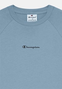 Lys blå bomulls t-skjorte med rund hals og raglanærmer, med en svart "Champion" logo på forsiden.