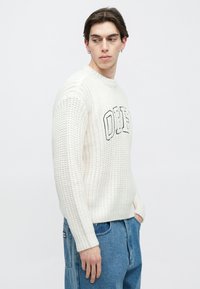 Pulover alb tricotat cu textură groasă și decolteu rotund. Prezintă broderia "OHE" în negru pe față. Asortat cu blugi din denim albastru.