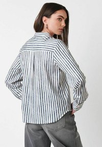 Frau mit langen dunklen Haaren, die silberne Creolen, ein weiß- und blaugestreiftes Langarmhemd und graue Hose trägt und über die Schulter schaut.