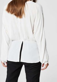 Weiße Bluse mit langen Ärmeln, gerafftem Rückendetail und offenem Schlitz an der Taille. Hergestellt aus leichtem, weichem Stoff mit glatter Textur.