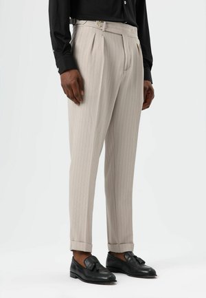 Pantalon beige à rayures fines avec un devant plissé, agrémenté de détails à boutons à la taille. Porté avec des mocassins noirs, mettant en valeur un ourlet retroussé.