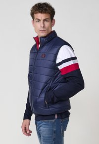 Chaleco puffy de color navy con cuello alto, diseño acolchado, cierre frontal y bolsillos con cremallera. Combinado con una camiseta de manga larga de bloques de color debajo.