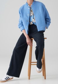 Veste bomber en laine bleu clair avec des boutons-pression, associée à une chemise à motifs, un pantalon large noir et des baskets blanches avec des accents gris.