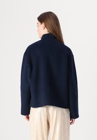 Marineblauer Wollmantel mit Stehkragen und abgesenkten Schultern. Verfügt über einen runden Akzent auf der Rückseite und einen lockeren Schnitt.