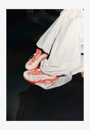 Fehér atlétikai cipők korall színű részletekkel és hálós felsőrésszel, vastag talppal és látható légbetéttel, világos színű anyagból készült nadrággal viselve.