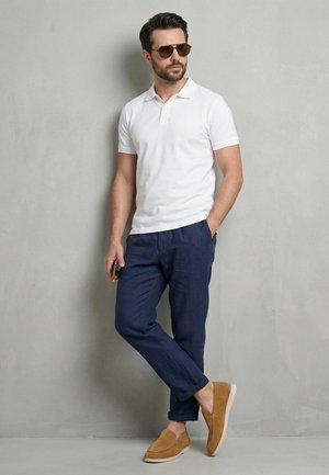 Mand iført hvid polo, blå bukser, beige loafers og solbriller, stående afslappet mod en grå væg, holder en telefon i højre hånd.