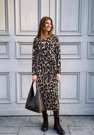 Une femme souriante, vêtue d’une robe midi à motifs et de bottes noires, porte un grand sac à main noir devant un mur clair à panneaux.