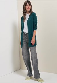 Tyrkysový cardigan s dlouhými rukávy a předními kapsami, nošený přes bílý top, spárovaný se šedými džíny a světle zelenými teniskami. Jednoduchý design, měkká textura.