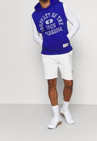 Hoodie senza maniche blu royal con una maglietta a maniche lunghe bianca, pantaloni corti bianchi e sneakers bianche. Il hoodie presenta un testo in grassetto e una tasca.