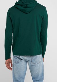 Mörkt grön långärmad hoodie med dragband i huvan, tillverkad av mjukt tyg. Har en avslappnad passform och tryckt etikett vid nederkanten.