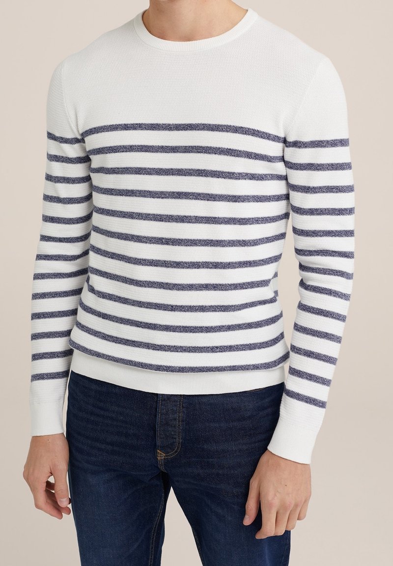 Homme portant un pull blanc à manches longues avec des rayures horizontales bleu marine assorti à un jean bleu foncé, debout devant un fond beige.