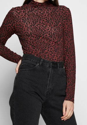Femme portant un haut à manches longues rouge et noir à imprimé léopard rentré dans un jean noir taille haute, main posée sur la hanche.