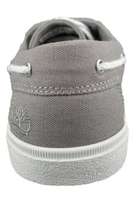Zapato de lona gris con logo bordado, ojales blancos y suela de goma texturizada blanca. La vista trasera se centra en el talón y el material superior.