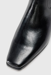Zapato de cuero pulido negro con punta cuadrada y textura suave sobre un fondo blanco.