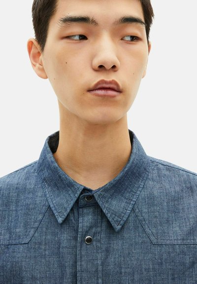 Camisa de chambray azul escuro com colarinho pontiagudo, fecho de botão e textura subtil. Apresenta yokes nos ombros e botões pretos em contraste.