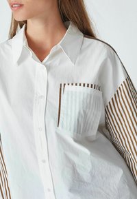 Camicia bianca con un tessuto liscio. Presenta una tasca marrone a righe e inserti marroni a righe sulle maniche. Chiusura con bottoni visibile.