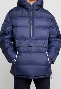 Granatowa kurtka puchowa z zamkiem z przodu, elastycznymi mankietami i bocznymi kieszeniami. Posiada ściągacz u dołu oraz dużą kaptur dla ciepła.