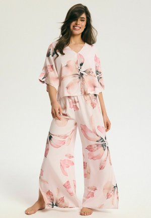 Kobieta stojąca boso, ubrana w luźny, jasnoróżowy dopasowany top i spodnie z szerokimi nogawkami z dużymi kwiatowymi wzorami, uśmiechająca się z długimi falującymi włosami.