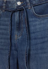 Denim-farkut keskisinisellä pesulla. Niissä on etutaskut, kontrastipistely, metallinen nappi sekä vyötärölle sidottava nauha.