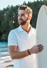 White polo shirt, beaded bracelet, holding a surfboard; sunlit beach, ocean waves in background, trees in distance.