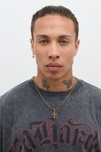 Svart, vintage-stil T-skjorte med røde bokstaver; gullkorshalskjede; subtile tatoveringer på nakken; kort, slicked-back frisyre.