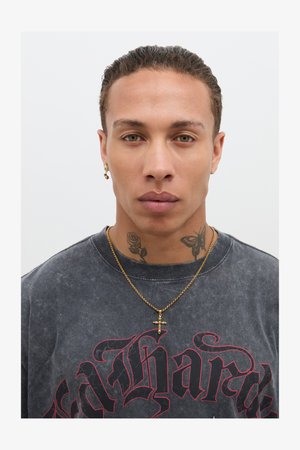 Black, vintage-style T-shirt with red lettering; gold cross necklace; subtle tattoos on neck; short, slicked-back hairstyle.