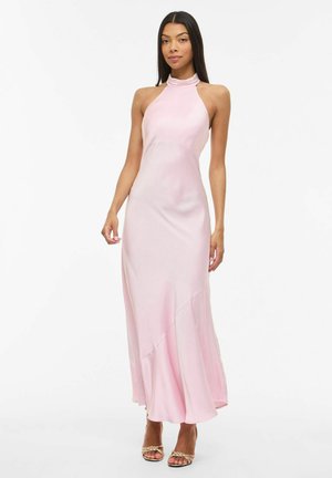Kvinne iført en ermeløs, lyserosa sateng kjole med halterhals og gullfargede åpne tå-sko, stående mot en hvit bakgrunn.