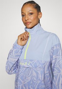 Jasnoniebieska kurtka przeciwwiatrowa z wysokim kołnierzem, z kontrastowym żółtym zamkiem błyskawicznym i kwiatowym wzorem. Logo "ROXY" w kolorze żółtym na klatce piersiowej.