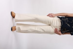 Femme debout portant un pantalon beige à jambes larges, un haut noir à motifs floraux et des mocassins marron clair sur un fond blanc uni.