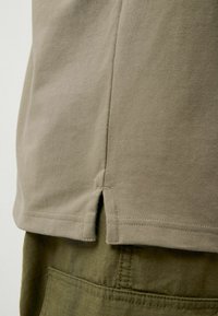 T-shirt in cotone khaki con fessure laterali, maniche corte e una texture liscia. Abbinato a pantaloni verdi con texture e dettagli di cucitura visibili.