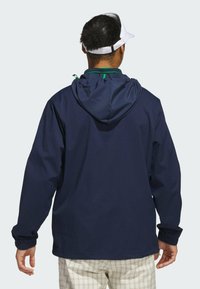 Chaqueta con capucha de color azul marino, con puños elásticos, capucha ajustable y detalles en verde. La vista trasera resalta un diseño simple y un tejido ligero.