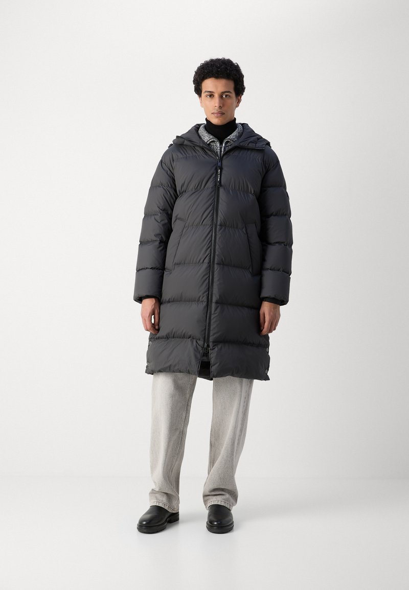 Abrigo acolchado negro con capucha, que presenta un cierre de cremallera en la parte delantera y un diseño acolchado horizontal. Llevado sobre pantalones gris claro, con un estilo minimalista.