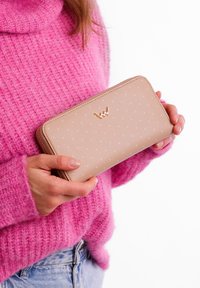 Beige wallet with polka dot pattern, zip closure, and gold logo detail. Held against a textured pink sweater.