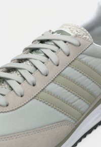 Αθλητικό παπούτσι με γκρι ανοιχτό και μπεζ επάνω μέρος, που διαθέτει τρεις ανοιχτοπράσινες ρίγες, επενδυμένο κολάρο και υφασμένα κορδόνια. Λαστιχένια σόλα.