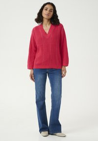 Pull en tricot rose avec un col en V, présentant une texture côtelée verticale et des détails de points ouverts, associé à un jean évasé bleu et des chaussures beiges.
