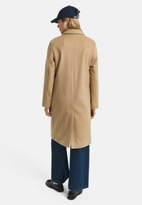 Beige wool blend coat with a back vent, notched collar, and dropped shoulders, paired with wide-leg dark denim pants and black shoes.