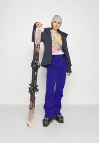 Skioutfit met een marineblauwe jas, een multicolor tie-dye shirt en blauwe skibroeken. Het model houdt een ski vast met zwarte bindingen en draagt een beige muts.