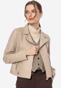 Beige leren jasje met een kraag, ritssluiting, zijzakken en een strakke vormgeving. Het model draagt een aansluitende crèmekleurige coltrui en een gebreid vest eronder.