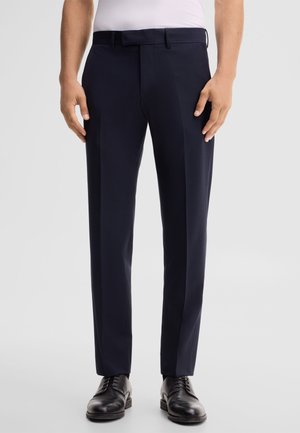 Uomo che indossa pantaloni eleganti blu scuro con piega frontale, scarpe eleganti nere e camicia bianca, in piedi davanti a uno sfondo chiaro e uniforme.