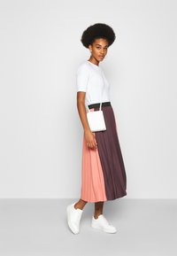 Bílý top s krátkým rukávem v kombinaci s plisovanou sukní s barevnými bloky v růžové, fialové a černé; doplněno malou bílou crossbody taškou a bílými teniskami.