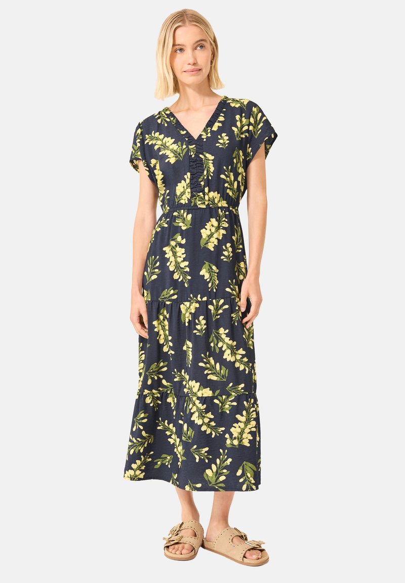 Femme aux cheveux blonds courts portant une robe midi bleu marine avec un imprimé floral jaune et des sandales beiges à enfiler, debout devant un fond uni.