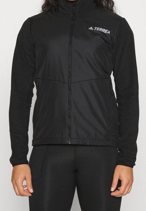 Personne portant une veste zippée Adidas Terrex noire et des leggings noirs, debout avec les bras détendus le long du corps.