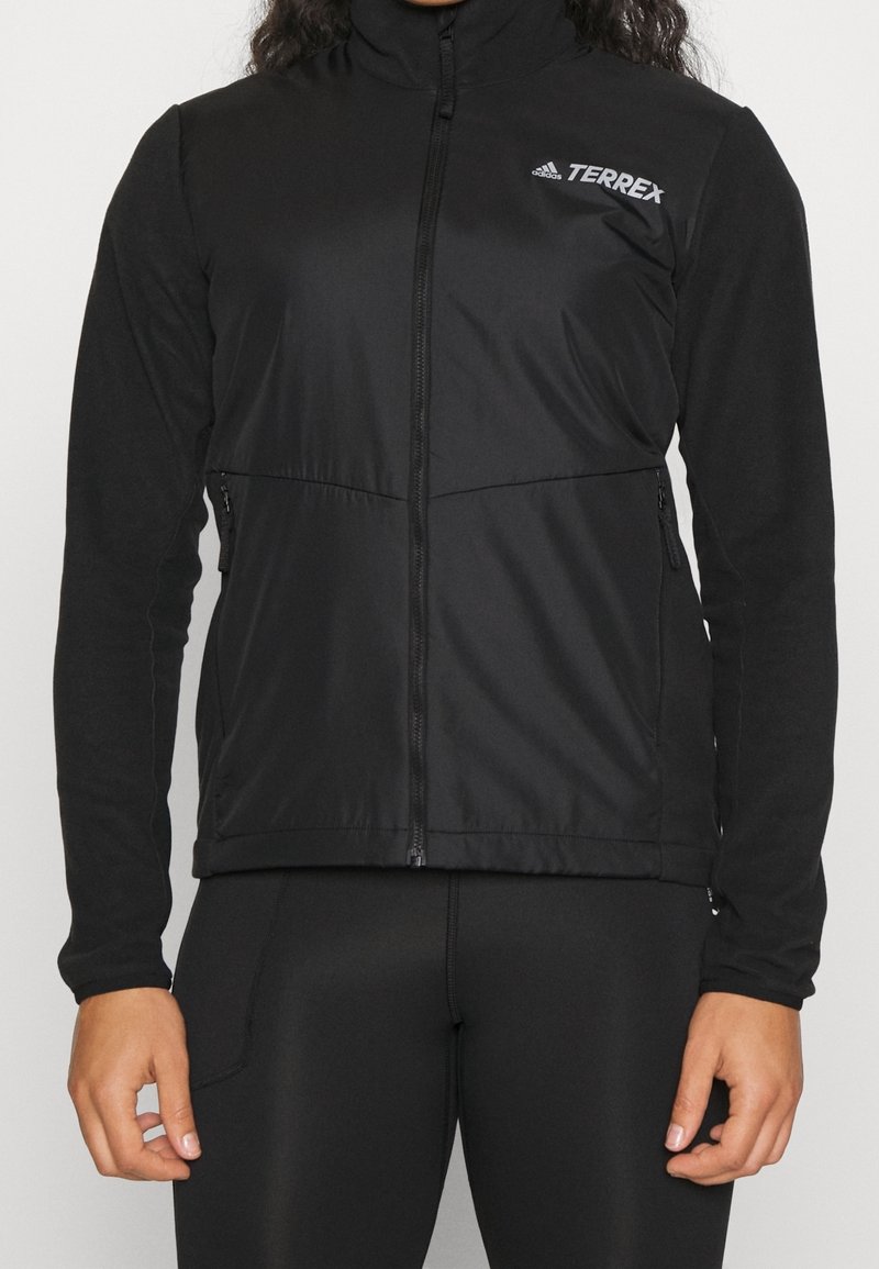 Personne portant une veste zippée Adidas Terrex noire et des leggings noirs, debout avec les bras détendus le long du corps.
