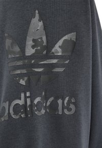 Sudadera gris oscura con un logo de Adidas en camuflaje negro texturizado que presenta tres formas de hoja y letras gruesas debajo del logo.