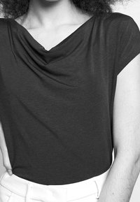 Kvinna som bär en svart kortärmad topp med draperad halsringning och vita byxor, sedd från axlarna till midjan.