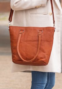 Borsa a tracolla in pelle marrone con due manici robusti, texture liscia e dettagli di cucitura minimi; portata sulla spalla di una persona.