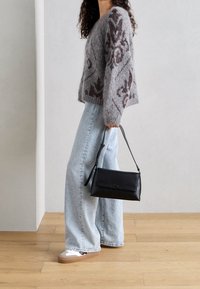 Žena v šedém vzorovaném svetru, světle modrých širokých kalhotách, bílých teniskách, držící černou kabelku přes rameno uvnitř na dřevěné podlaze.