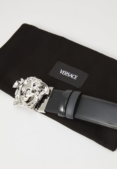 Curea neagră cu o cataramă în formă de cap de leu argintie, textură netedă, însoțită de un pouch din material textil negru cu eticheta albă "VERSACE".