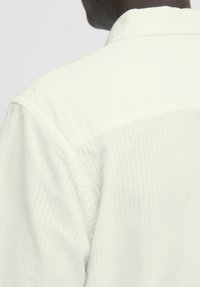 Camisa blanca con una superficie texturizada, que presenta un collar estándar y sutiles rayas verticales. La parte trasera está diseñada con un acabado suave.